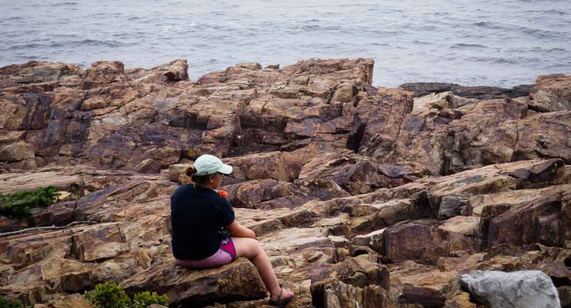 a person sits on a rock along a rocky shoreline while on an outward bound course in maine